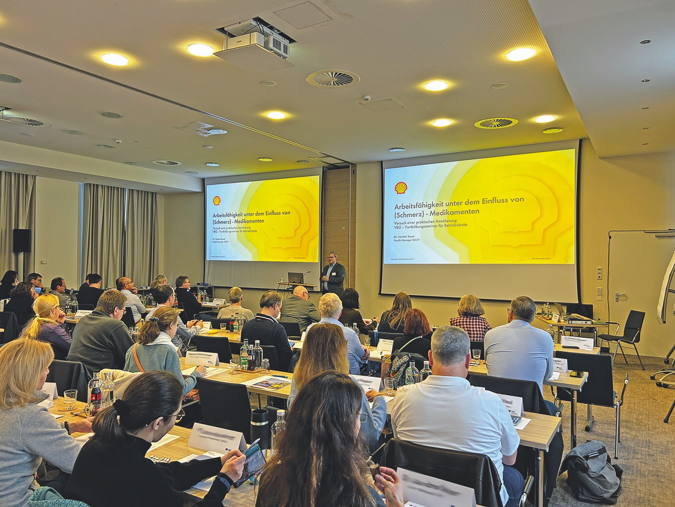 Mit Teilnehmern gefüllter Seminarsaal vor zwei großen Bildschirmen